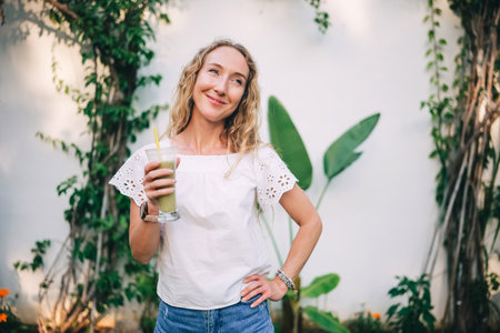 A smiling woman with wavy hair holds a green beverage while standing near tropical plants.の写真素材