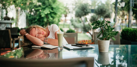 A woman relaxes with her head resting on a notebook in a vibrant cafe surrounded by greenery.の写真素材