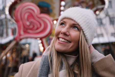 Portrait of young smiling woman wearing beige coat, white knitted hat and wide scarf holding heart shape lollipop in winter time outdoors. Christmas holidays.の写真素材