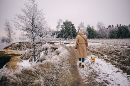 Happy attractive young woman wearing beige coat, white hat and mittens walking with welsh corgi pembroke puppy, outdoors near winter sea. Backside viewの写真素材