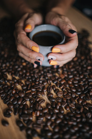 Hands with bright nails holding mug of coffee, set against a backdrop of coffee beans.の写真素材