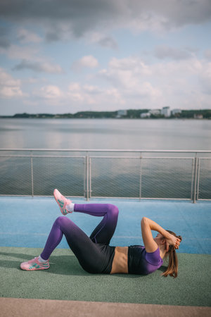 A woman exercises outdoors by a body of water, doing crunches on a fitness mat in activewear.