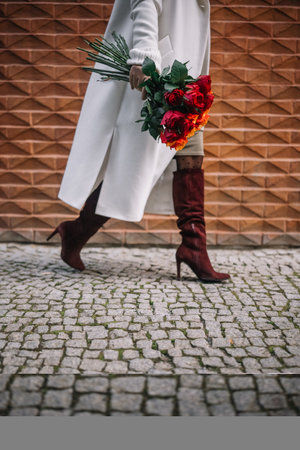 A woman in a long coat strides confidently on a cobblestone path, holding a bouquet of flowers.の写真素材