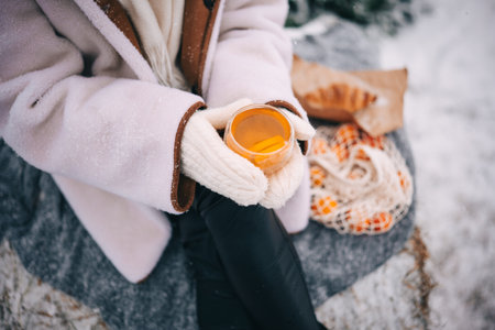 Person holding a warm drink, dressed in cozy winter attire, surrounded by a snowy landscape.の写真素材