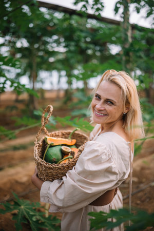 A woman smiles while holding a basket of fruits in a vibrant garden filled with greenery.