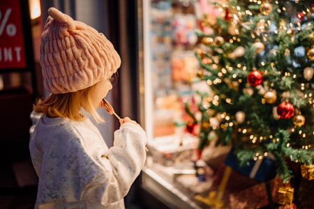 Young girl in a cozy hat admires a festive Christmas tree while savoring a lollipop outdoors.の写真素材