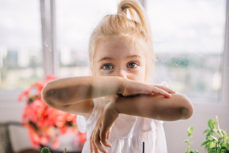 A girl with blonde hair smiles and playfully rests her arms on a glass window, surrounded by greenery.の写真素材
