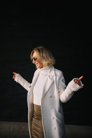 A woman dressed in a white coat and sunglasses poses playfully with her arms outstretched.の写真素材