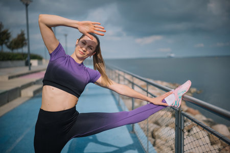 A woman stretches her leg by the water, wearing purple athletic attire on a cool, cloudy day.