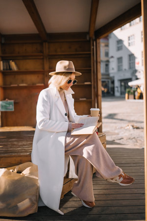 Relaxed atmosphere as a woman sips coffee and reads a book in an urban setting with wooden benches.の写真素材