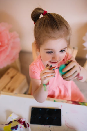 Cute happy 3 years old girl wearing pink dress painting Easter eggs in bright colourful roomの写真素材