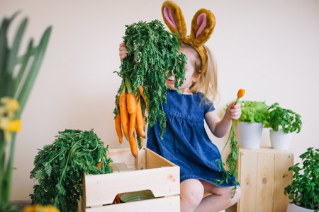 Cute little child girl wearing bunny ears sitting around wooden boxes with flowers and fresh vegetables and holding fresh bunch of carrotsの写真素材