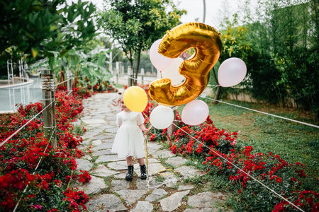 A child wearing a white dress holds balloons and stands among vibrant flowers in a garden pathway.の写真素材