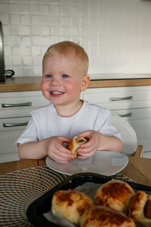 A cheerful toddler is seated at a dining table, holding a pastry with a big smile, surrounded by delicious baked goods.の写真素材