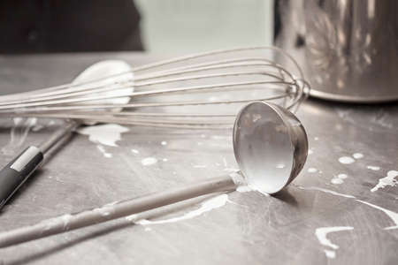 Close up of Inox steel kitchen surface, ladle and whisk tainted with spilled milk cream - Commercial...