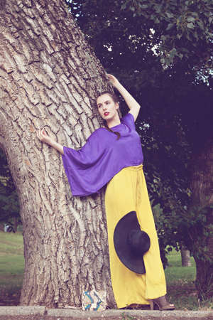 Pretty young woman fashion model posing for camera while standing by the tree in the parkの写真素材
