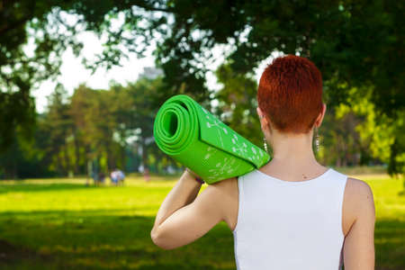 Exercise in nature, recreation in beautiful sunlit park. Beautiful female fitness instructor holding an exercise mat above her arm, active sport lifestyleの写真素材
