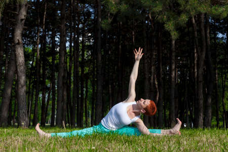 Exercise in nature-Redhead girl doing Yoga in the park, pine trees behind, sunlit, horizontalの写真素材