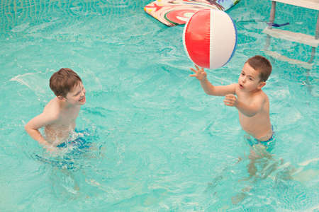 Happy kids playing in the swimming pool with ball and water mattress. Having fun in water pool outside on summer day. Leisure and swimming at holidays. Happiness and joy.の写真素材