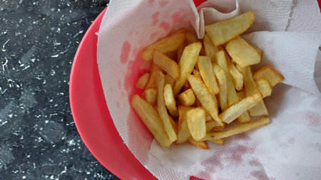 Golden French fries in a red plate on a black granite table.