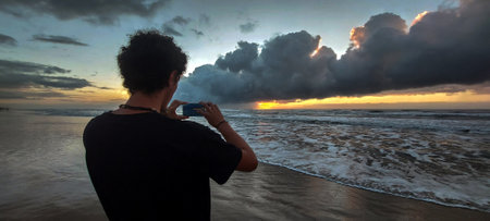 Young man taking a photo of the sunset on the beach of Bali