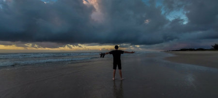 A man in a black T-shirt and shorts stands on the sand and looks at the sea.の写真素材