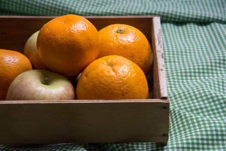 fresh oranges and apples in a wooden box on a green tableclothの写真素材