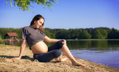 Young pregnant woman in a sport dress sitting on beach near lake