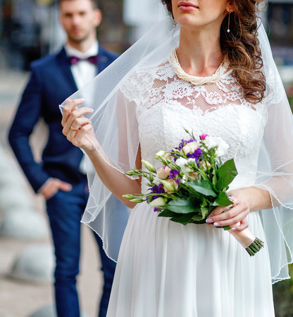 bride in lace veil holding a wedding bouquet. the groom in a tuxedo in the backgroundの写真素材