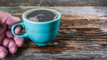 A hand holding green mug of coffee sits on a wooden table, close upの写真素材