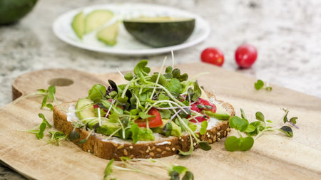 A delicious open-faced sandwich is displayed on a wooden board, featuring a generous layer of avocado, sliced tomatoes, and an abundance of vibrant microgreensの写真素材