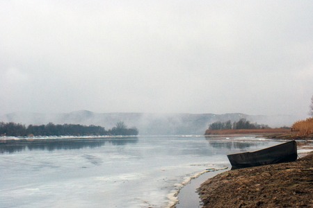 Old boat on the river bank covered with ice and fog in winterの写真素材