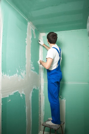 Portrait of young pretty man on construction site: male worker plastering walls with spatula while remodeling officeの写真素材