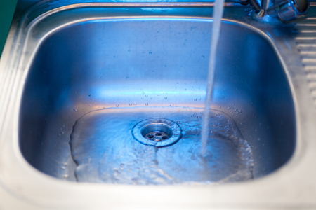 A stream of clean, transparent fresh water flows from the steel shiny tap into a stainless steel sink in blue tones. Water drains into the hole for the shell close-up macroの写真素材