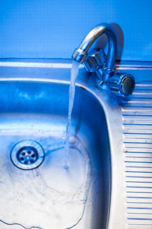 A stream of clean, transparent fresh water flows from the steel shiny tap into a stainless steel sink in blue tones. Water drains into the hole for the shell close-up macroの写真素材