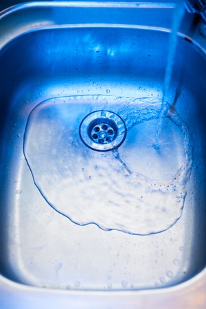 A stream of clean, transparent fresh water flows from the steel shiny tap into a stainless steel sink in blue tones. Water drains into the hole for the shell close-up macroの写真素材