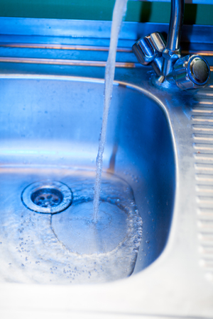 A stream of clean, transparent fresh water flows from the steel shiny tap into a stainless steel sink in blue tones. Water drains into the hole for the shell close-up macroの写真素材