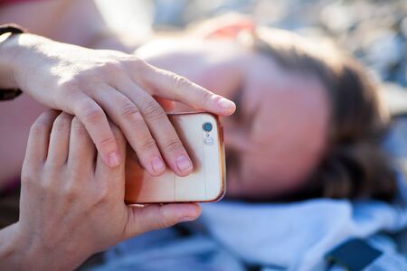 Pretty teenager girl using a smart phone lying on the beach with the sea and horizon in the...