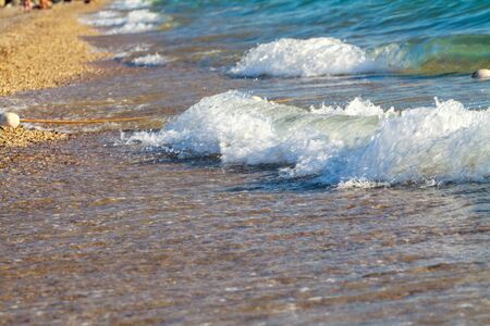 Sea waves with white foam on the seashore. Summer beach of the warm sea. The sea shore is made of shells and sand and is surrounded by salt waves with foam.の写真素材