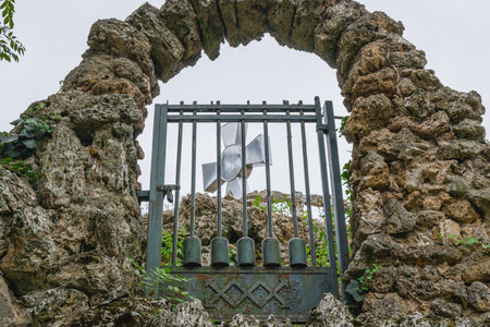 Old stone archway with a weathered metal gate and cross symbol, framed by rugged rocks and greenery,...