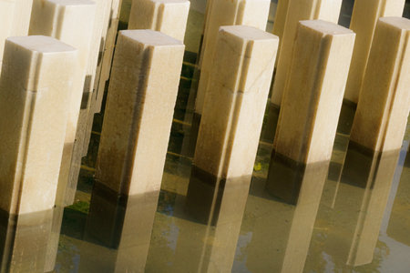 Close-up of vertical stone columns reflected in still water during the golden hour. The composition highlights the architectural geometry, surface texture, and serene interplay of light and reflection.の写真素材