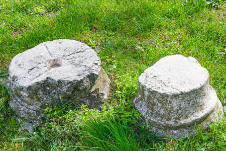 Two weathered stone column bases lie on a grassy field, illuminated by sunlight. The aged texture and moss growth highlight the passage of time and the preservation of ancient history.の写真素材