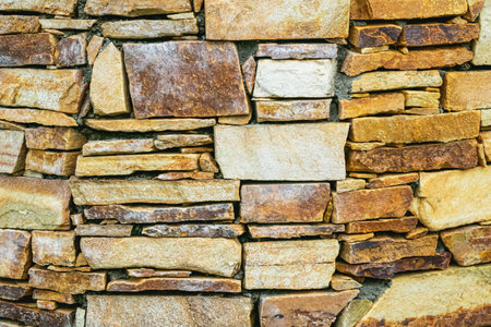 Close-up of a rustic stone wall built from uneven natural rocks in shades of brown, beige, and...