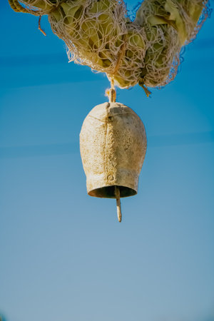 An old rustic bell made of weathered metal hangs suspended from a coarse rope under a bright blue...