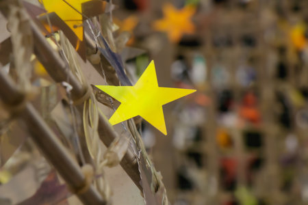 Close-up view of a bright yellow paper star tied to a decorative frame, part of a festive community display. The shallow depth of field highlights the starâs vivid color while soft background lights create a warm and joyful atmosphere.の写真素材