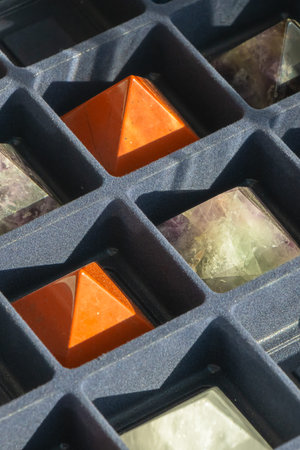 Close-up view of polished crystal pyramids placed in a foam display tray with geometric compartments. Sunlight highlights textures, colors, and mineral patterns in the stones.の写真素材