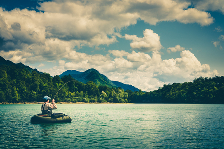 Fisherman sitting in bellyboat and fighting with big trout on lake in Jesenice, Slovenia. Still water fly fishing and outdoor lifestyle theme. Moody, contrasty, toned and faded look.の写真素材