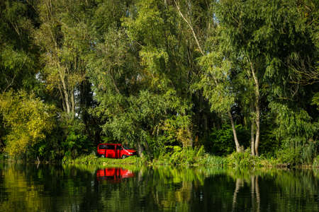 Red camper van on shore of Danube river with green trees and grass in background and with reflecion in water surface.の写真素材