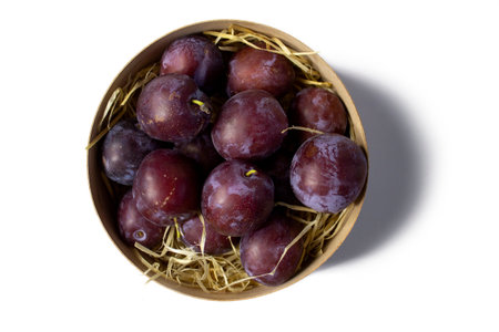 Plums in wooden basket and straw isolated on white background, top view