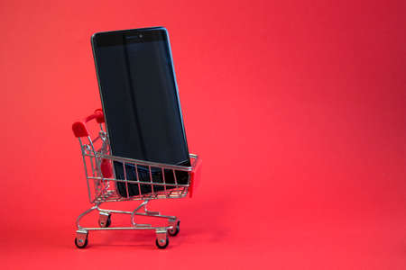 Modern black smartphone in small shopping cart from supermarket close up on bright red background with blank empty spaceの写真素材
